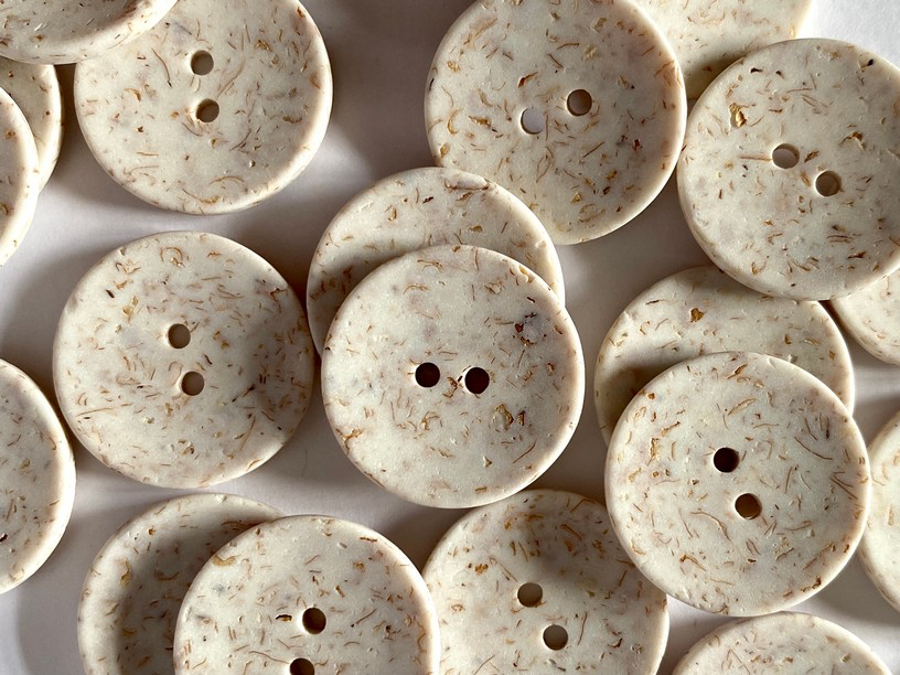 A close-up pile of round two-hole buttons made from a textured cream-colored material with fine tan and brown flecks, set against a matching light background.