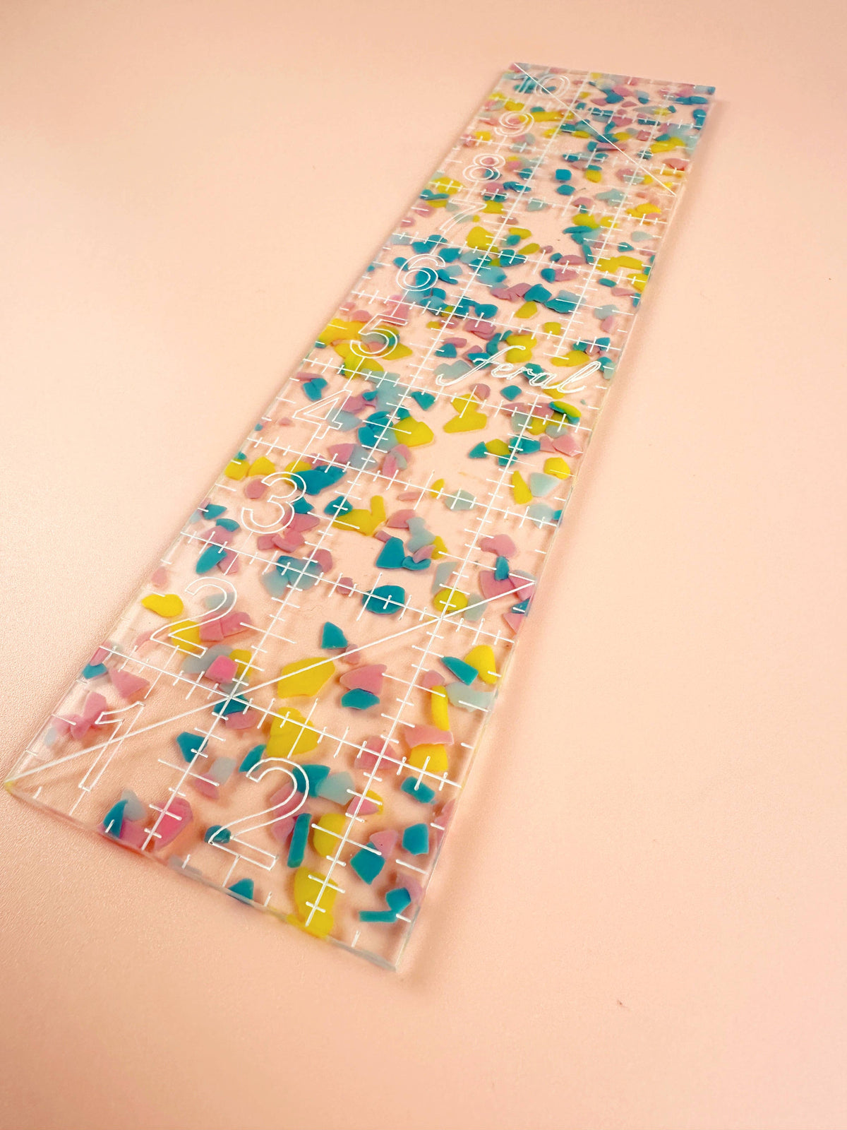 Feral Notions 2.5 x 10 inch quilting ruler in rainbow confetti acrylic angled on peach, a strip cutting ruler