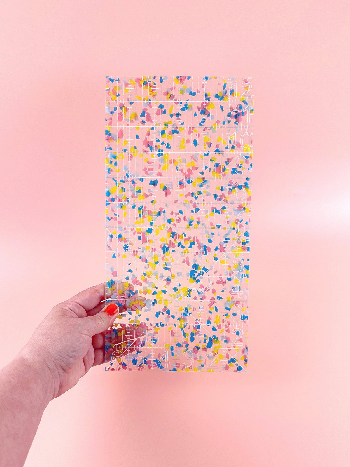 Hand holding the Feral Notions 6 x 12 inch quilting ruler in rainbow confetti acrylic