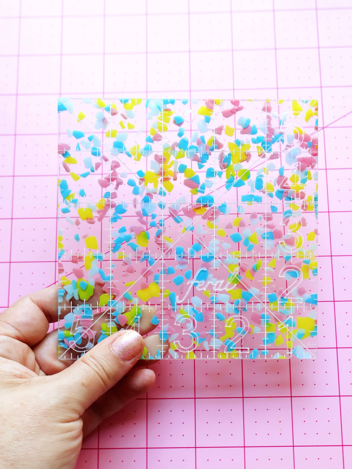 Hand holding the Feral Notions 5 inch charm square quilting ruler in rainbow confetti acrylic over a cutting mat
