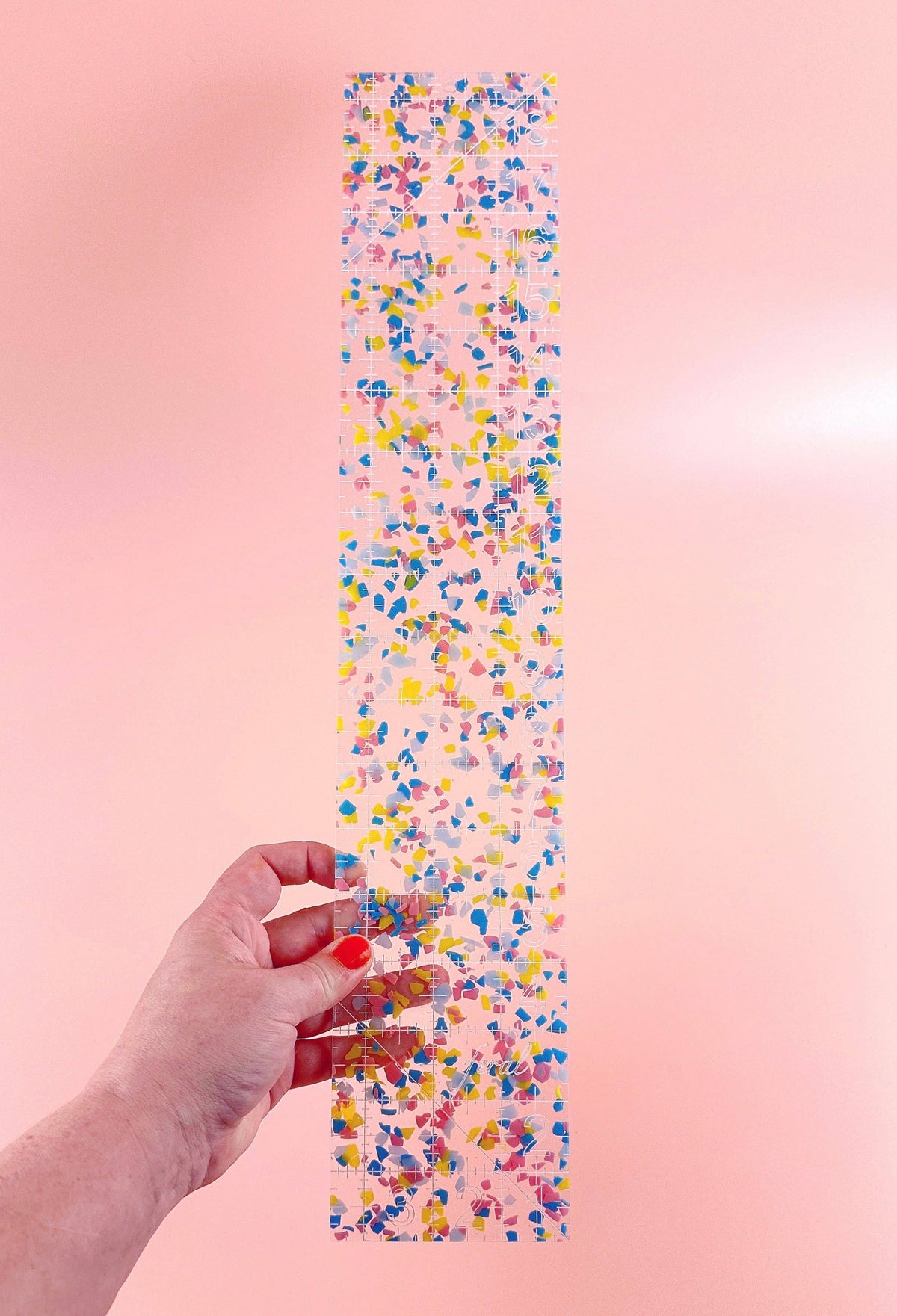 Feral Notions fat quarter quilting ruler in rainbow confetti acrylic held up in hand on a peach background