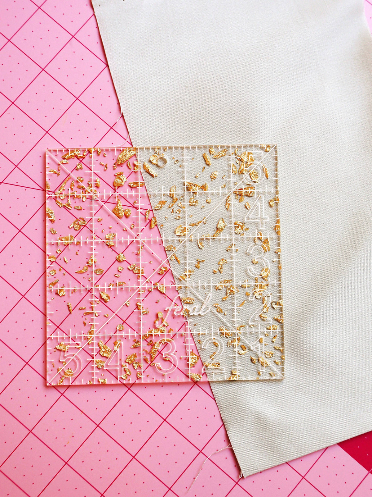 Feral Notions 5 inch charm square quilting ruler in gold glitter acrylic on white fabric over a cutting mat