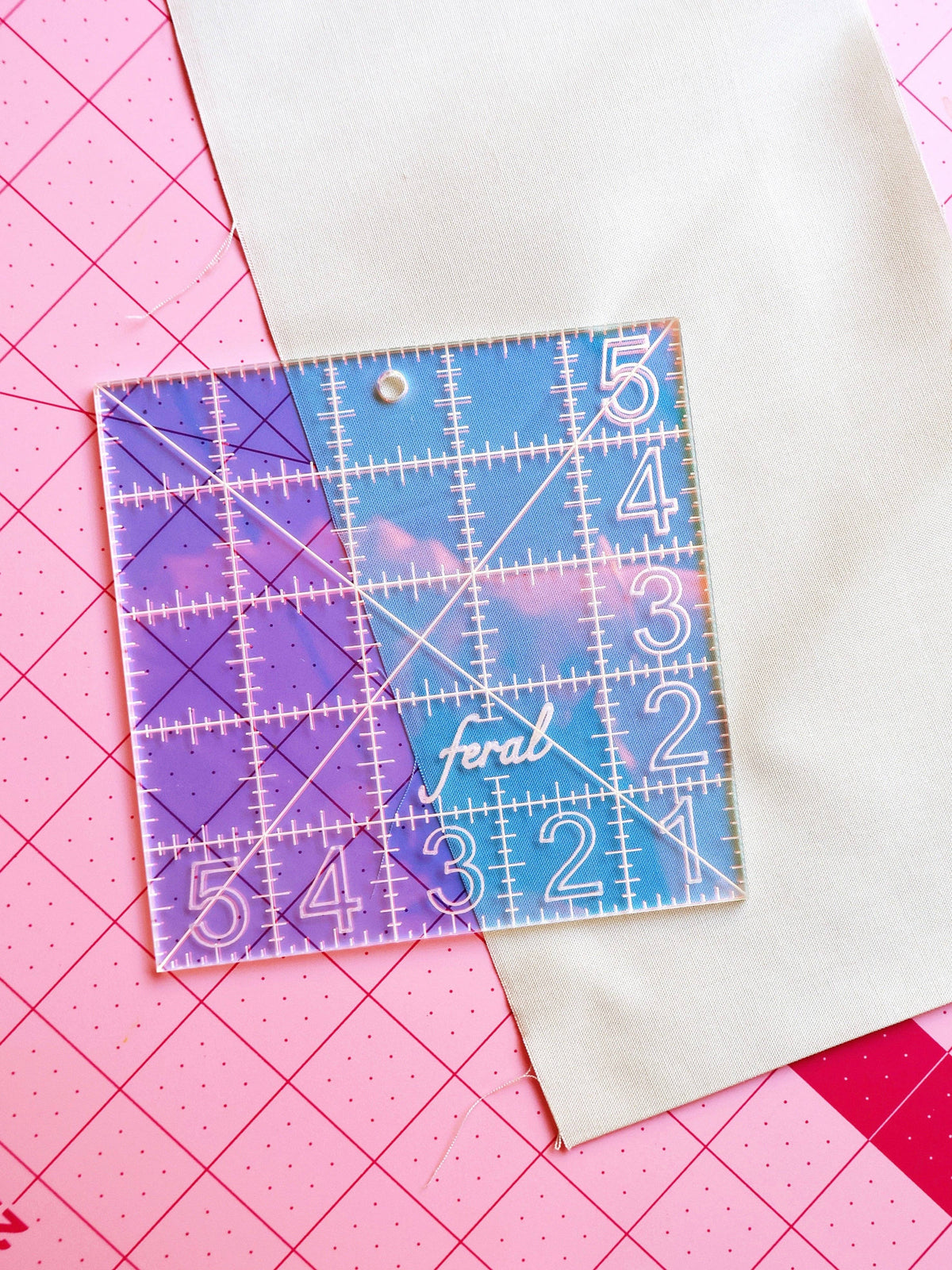 Feral Notions 5 inch charm square quilting ruler in iridescent acrylic over white fabric on a cutting mat