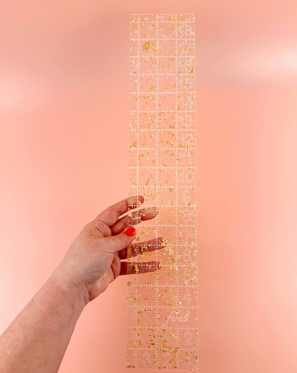 Hand holding the Feral Notions fat quarter quilting ruler in gold glitter acrylic to show the measuring lines