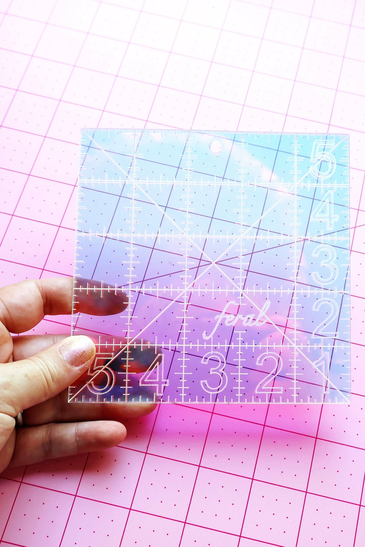 Feral Notions 5 inch charm square quilting ruler in pale iridescent acrylic on a pink cutting mat