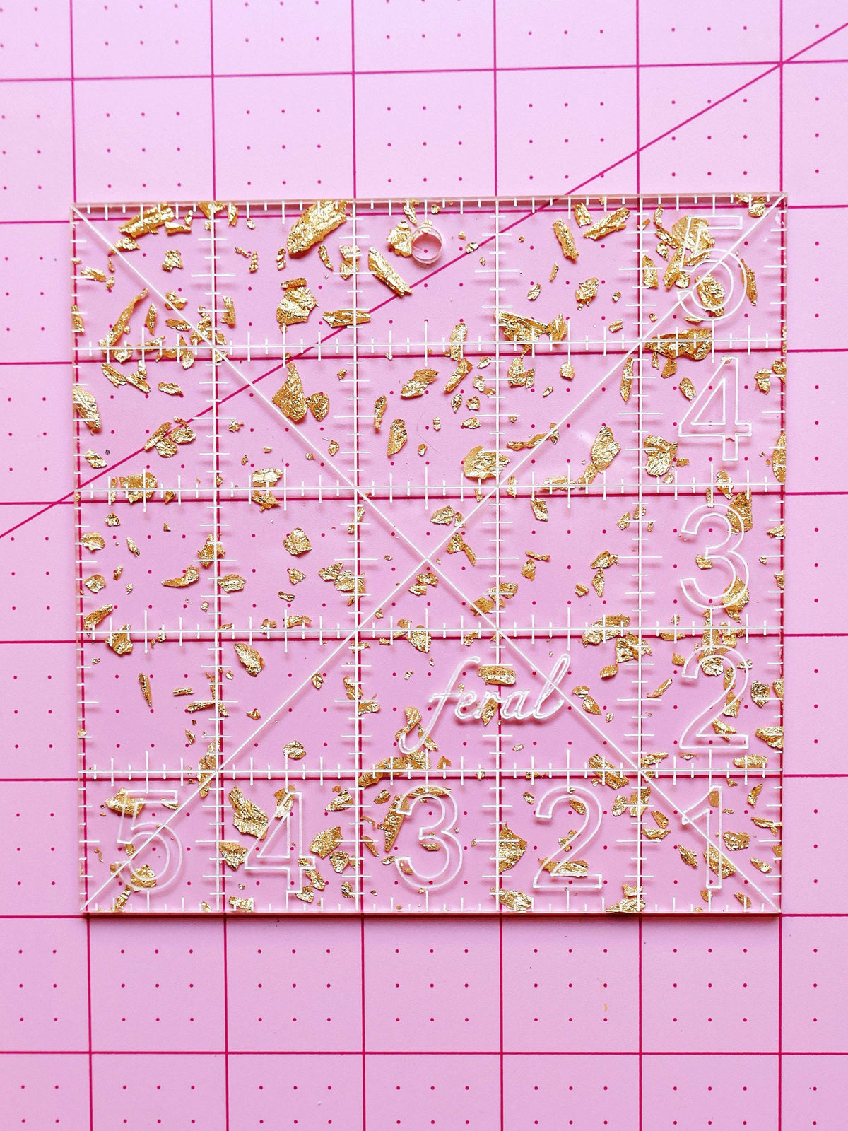 Feral Notions 5 inch charm square quilting ruler in gold glitter acrylic on a pink cutting mat