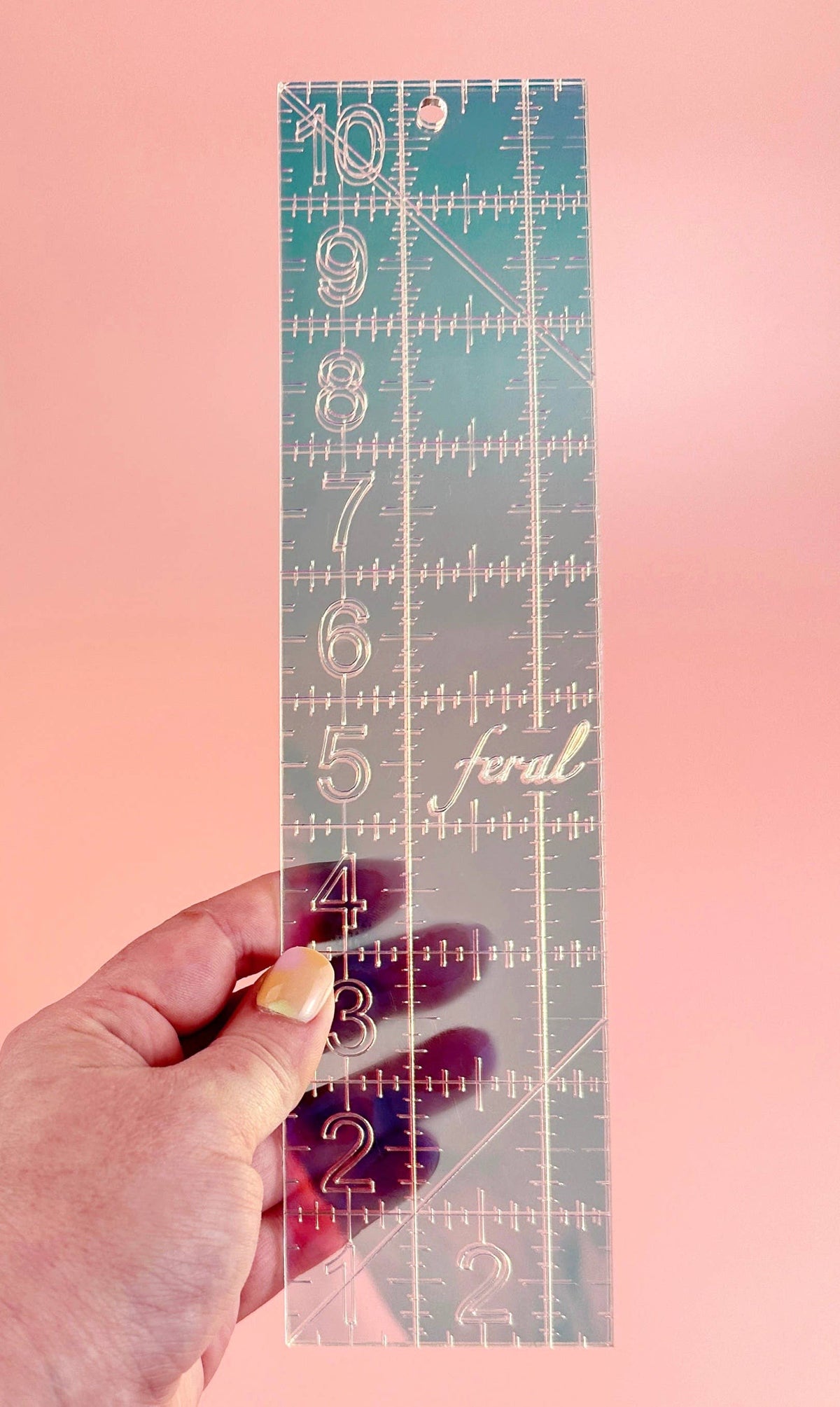 Hand holding the Feral Notions 2.5 x 10 inch quilting ruler in iridescent teal acrylic