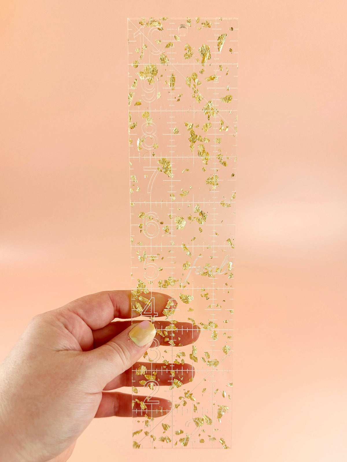 Hand holding the Feral Notions 2.5 x 10 inch quilting ruler in gold glitter acrylic