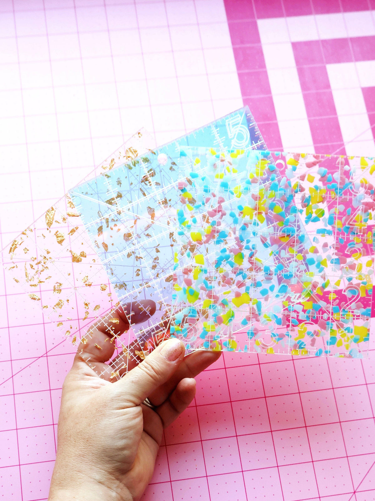 Hand holding the Feral Notions 5 inch charm square quilting ruler in confetti acrylic over an iridescent one