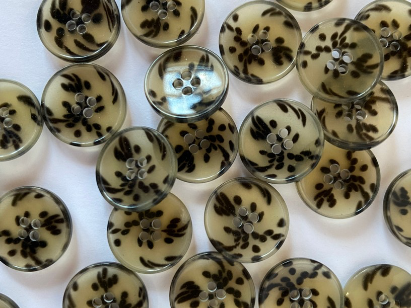 A close-up arrangement of round decorative buttons with a translucent beige base and black floral or leaf-like patterns, photographed on a white background.