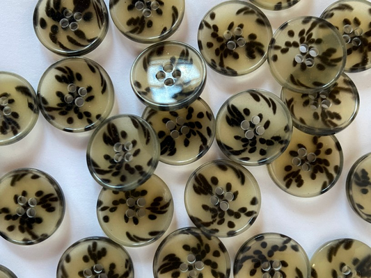 A close-up arrangement of round decorative buttons with a translucent beige base and black floral or leaf-like patterns, photographed on a white background.