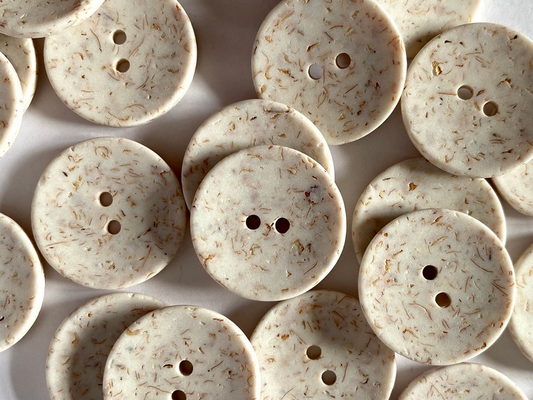 A close-up pile of round two-hole buttons made from a textured cream-colored material with fine tan and brown flecks, set against a matching light background.
