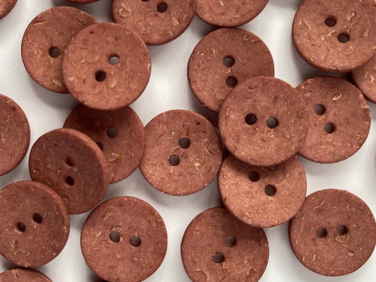 A close-up pile of round, two-hole buttons made from a textured brown material, possibly cork or compressed wood, scattered on a white background.