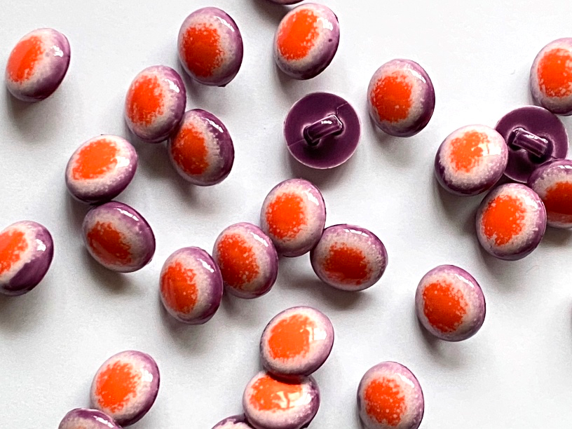 A close-up of multiple small oval buttons scattered on a plain white background. Each button has a glossy purple outer edge and a bright orange center with a white marbled or speckled transition around the perimeter.
