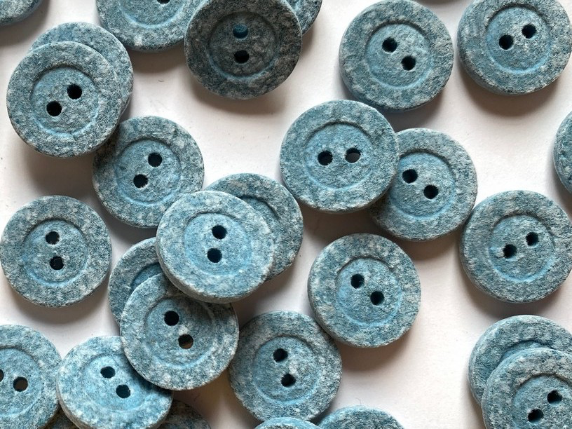 A pile of round, two-hole buttons with a textured, stone-like finish in a dusty slate blue color, scattered on a plain white background.