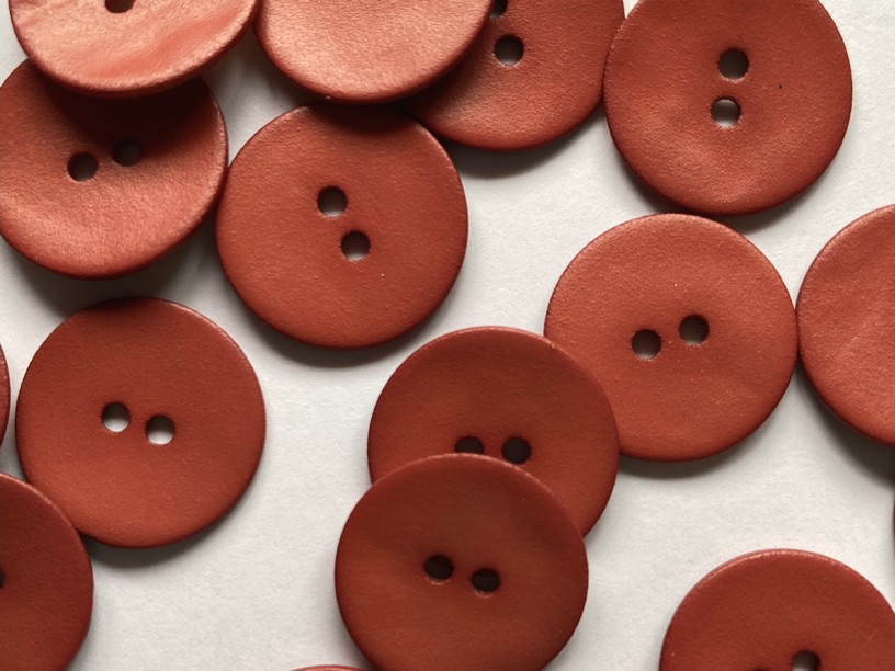A close-up of multiple round, two-hole buttons in a warm terracotta or rust red color scattered on a plain white background.