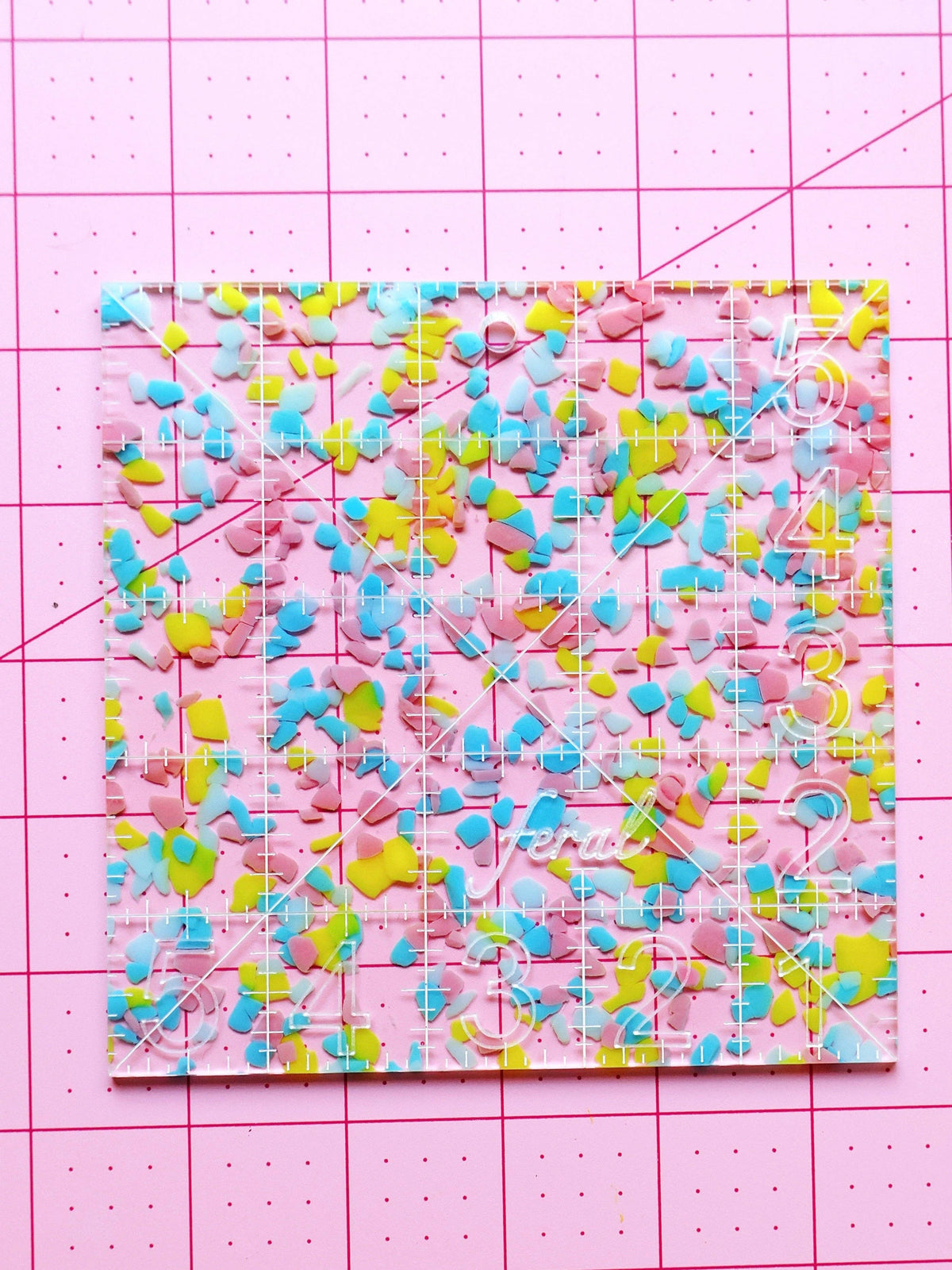 Feral Notions 5 inch charm square quilting ruler in rainbow confetti acrylic laid flat on a cutting mat