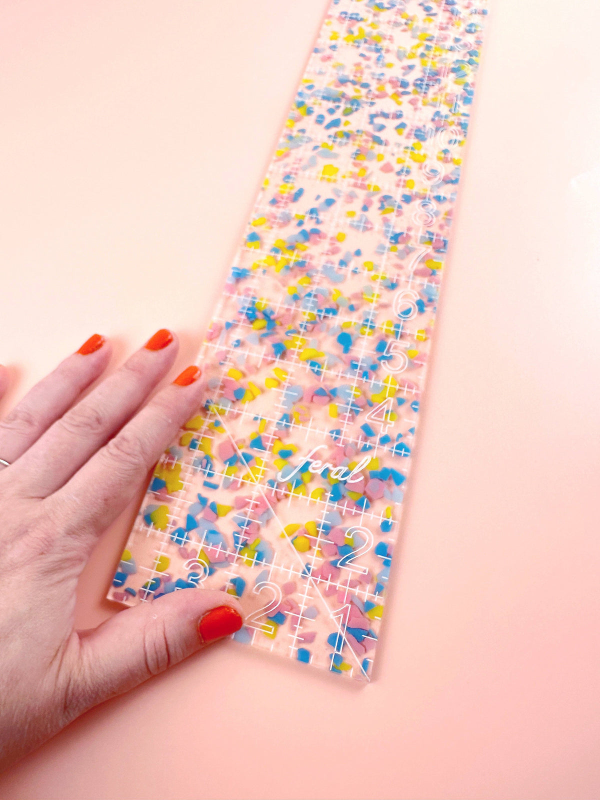 Hand holding the Feral Notions fat quarter quilting ruler in rainbow confetti acrylic, close-up of the speckles