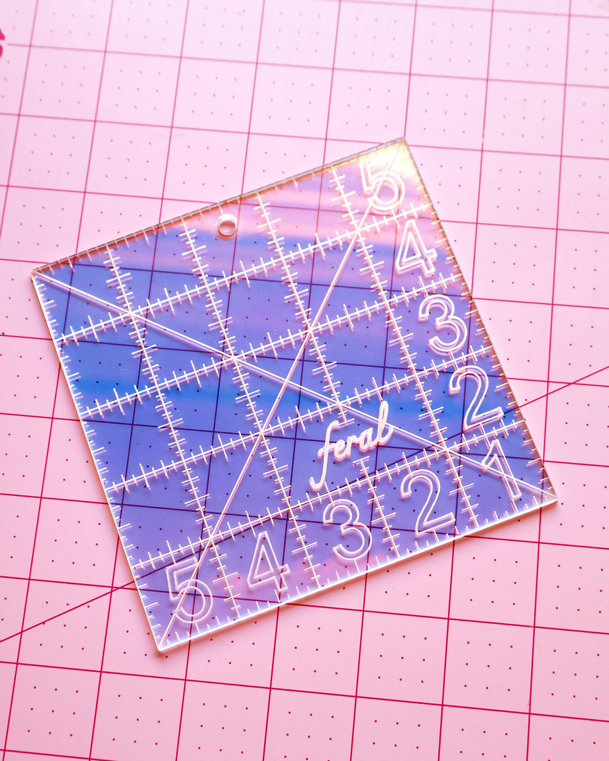 Feral Notions 5 inch charm square quilting ruler in clear iridescent acrylic on a pink cutting mat