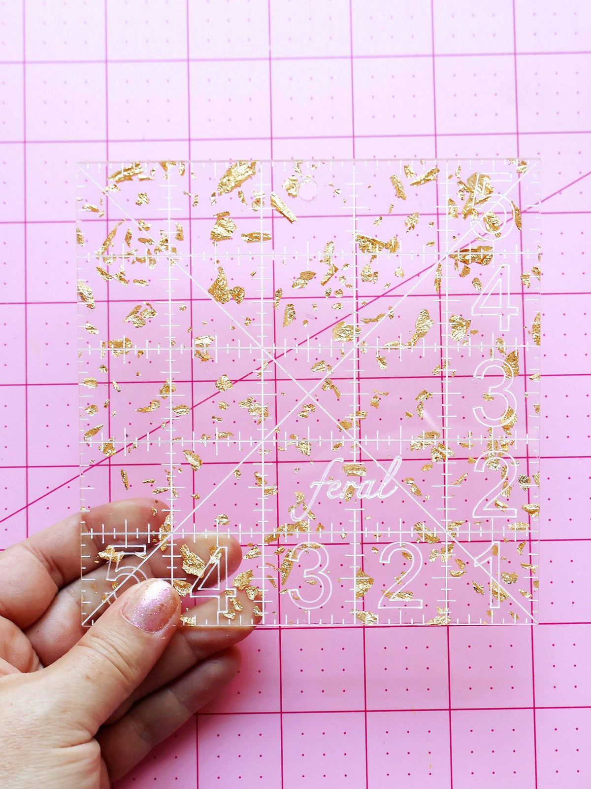 Hand holding the Feral Notions 5 inch charm square quilting ruler in gold glitter acrylic on a cutting mat