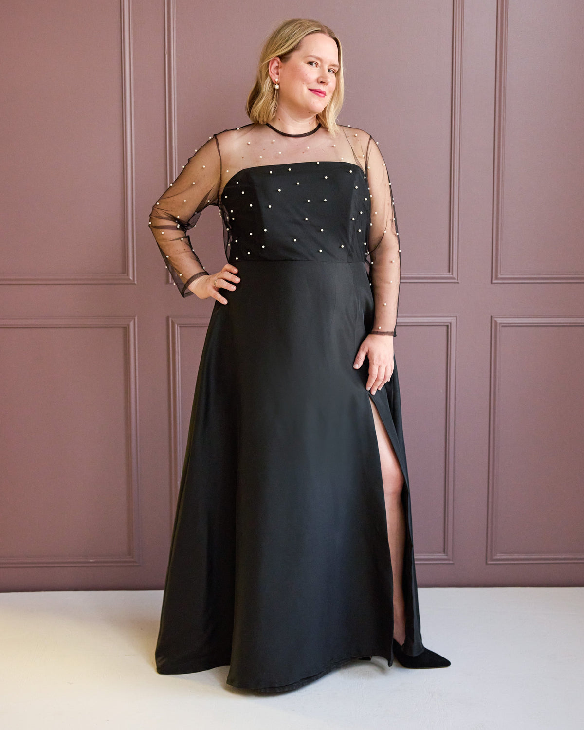 Cashmerette Ellington Dress PDF sewing pattern, woman wearing a black evening gown with sheer sleeves against a pink paneled wall, side view