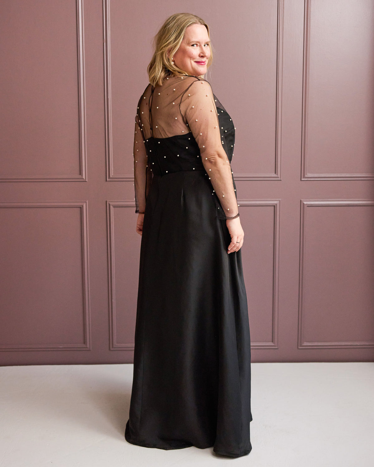 Cashmerette Ellington Dress PDF sewing pattern, woman wearing a black evening gown with sheer sleeves against a pink paneled wall, back view