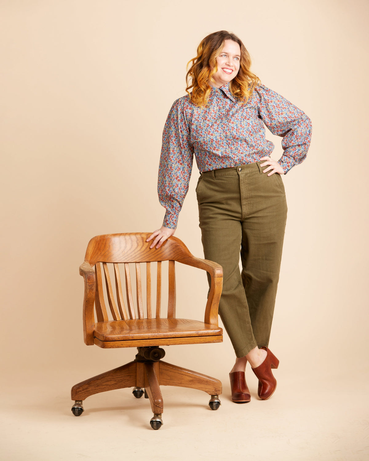 Cashmerette Vernon Shirt PDF sewing pattern, woman in a patterned blouse and green pants standing next to a wooden chair